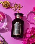 A beautiful display featuring a jar of Symbeeosis: Beautifying Immunitea for the Queen Bee on a vibrant pink background. Surrounding the jar are a transparent teapot, two transparent cups, a spoon with loose leaf tea, and assorted colorful flowers. This serene setup invites you to start your wellness journey with health-boosting ingredients.