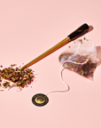 A gold and black spoon holds the Venus – Beauty Sleep Herbal Tea Bags’ botanical herbal blend spilling beside a pyramid tea bag with a round "Magic Hour" label, all set on a pink background.