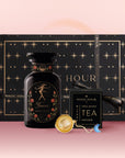 The Taurus Astrology Tea Gift Set includes a black and gold box, "Taurus Magic Hour" bottle, "Opal Moon Ritual Tea" packet, and gold tea infuser—ideal for astrology tea lovers. Beautifully arranged on a pink surface.