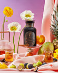 A black bottle labeled "Taurus Magic Hour" sits on a gold-stand, surrounded by sliced figs, a pineapple, a diced mango, and glass jars of liquid. Bright yellow and white flowers are in the background, with a pastel pink to purple gradient backdrop and draped fabric.