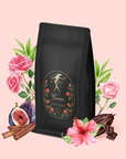 A bag of Taurus: Tea of Venusian Garden Delights with a decorative label is set among roses, hibiscus, figs, cinnamon sticks, cocoa pods, coffee leaves, and chocolate pieces on a pale pink background.