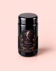 A black cylindrical container labeled “Taurus: Tea of Venusian Garden Delights,” adorned with an illustration of a woman, stars, and roses, is set against a soft pink background. This luxurious tea blend offers antioxidant-rich ingredients for a refined and healthful experience.