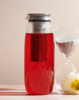 A Unitea Cold Brew Carafe filled with vibrant red liquid stands next to a delicate white flower and a clear hourglass with white sand. Featuring a built-in infuser basket, this carafe is an elegant piece of teaware. The neutral and minimalistic background highlights its modern design.