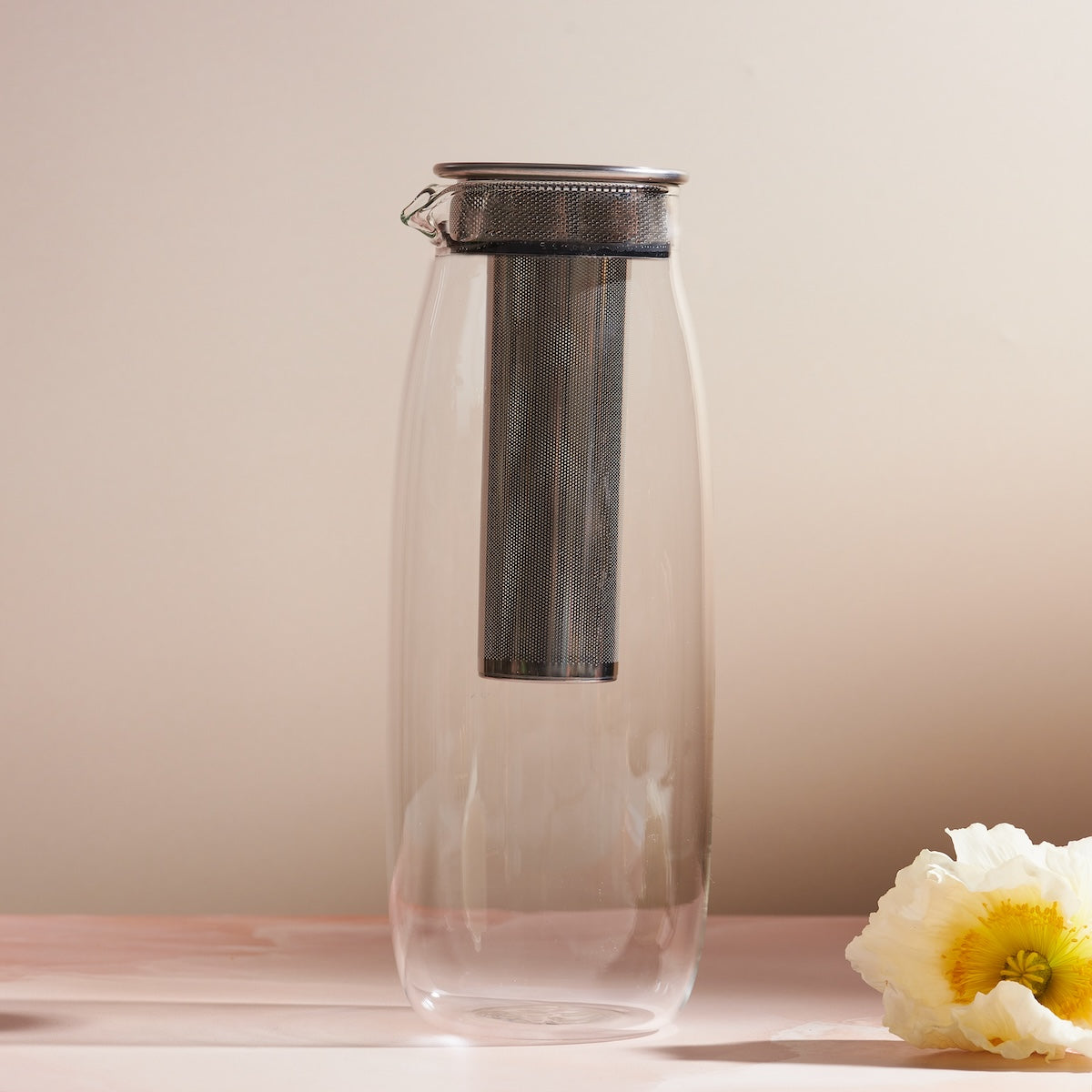 The Unitea Cold Brew Carafe, made of transparent glass and featuring a metal lid with a tall, cylindrical mesh infuser inside, rests against a neutral background. A white and yellow flower is placed to the right on the surface, enhancing the elegant teaware display.