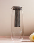 The Unitea Cold Brew Carafe, made of transparent glass and featuring a metal lid with a tall, cylindrical mesh infuser inside, rests against a neutral background. A white and yellow flower is placed to the right on the surface, enhancing the elegant teaware display.