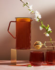 A still life composition featuring a tall Cast Sepia Pitcher, crafted from borosilicate glass, accompanied by two matching glasses and a brass tea infuser held by a hand. A delicate white blossom on a branch enhances the elegant and minimal aesthetic, set against a soft background.