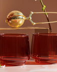 Two Sepia Tumblers from the Set of Four sit on a pink surface, their amber-colored glass evoking a vintage-inspired elegance. Above them, a gold tea infuser rests on a brass stand, with white petals scattered around. A delicate branch with small blossoms in the background adds to the warm and sophisticated scene.