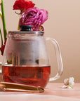 A Magic Hour Kinto Stainless Steel Teapot filled with organic tea sits on a table. The teapot has a stainless steel infuser inside and a clear lid with a black knob. Beside it is a pair of gold-colored tongs and a small white flower. In the background, there are vibrant pink and red flowers.