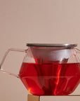 A Kinto Carat Teapot filled with rich red tea is shown against a beige background. The teapot, made of transparent glass, features a stainless steel lid and a handle on the left side. The vibrant color of the tea is beautifully visible through the clear glass.