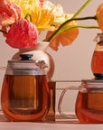 Two Magic Hour Kinto Stainless Steel Teapots filled with amber loose leaf tea are in the foreground, while a beige vase containing vibrant orange, pink, and yellow flowers is in the background. A third Magic Hour Kinto Stainless Steel Teapot is partially visible to the right. The setting is illuminated by soft light, evoking a magic hour tea experience.