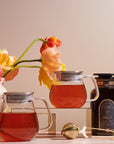 Two Kinto One Touch Teapots filled with organic tea are placed on a table, accompanied by a jar labeled "Magic Hour." A gold tea infuser and string lights lie in the foreground. In the background, there's a ceramic vase with colorful flowers and a soft, warm light illuminating the scene.