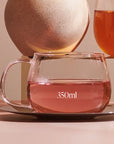 A clear Kinto Glass Tea Cup from Magic Hour, labeled "350ml," sits on a reflective saucer. Behind it, part of another glass filled with organic tea is visible, with a large, round stone in the background. The scene is set on a light pinkish-brown surface.