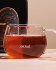 A Kinto Glass Tea Cup filled with 350ml of organic tea sits on a table against a neutral background. Behind the cup, there is a bottle labeled "Magic Hour - Timeless." The scene is well-lit, highlighting the tea's amber color.
