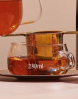 Close-up image of a 230ml Kinto Glass Tea Cup with a metal tea infuser placed on top. Organic tea is being poured from a glass teapot through the infuser into the cup. The setting is warm and inviting, perfect for preparing loose leaf tea during your Magic Hour tea ritual.