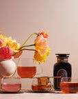 A still life image features a vase with colorful flowers, four Kinto Glass Tea Cups filled with different shades of organic tea, and a bottle of Magic Hour Tea. The background is softly lit with a warm tone, creating a cozy and inviting atmosphere.