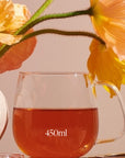 A Kinto Glass Tea Cup filled with a reddish beverage sits beside a white vase holding vibrant yellow and orange poppy flowers. The cup has "450ml" written on it. The soft lighting casts gentle shadows, creating a warm and serene atmosphere, perfect for savoring your Magic Hour tea.