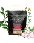 A black bag labeled "Small Batch Magic" features a pink teacup, tea leaves, herbs, and garlic. The Lapsang Souchong Turkey Brine is infused with smoked black tea, offering a rich, unique flavor for gourmet turkey dishes.