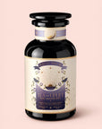 A dark glass bottle labeled "Tea to Uplift: Earl Greater Grey: Black & White Tea with Bergamot," adorned with gold and purple accents, floral illustrations, and a sunburst design, is displayed against a soft pale pink background. This sophisticated presentation showcases the exquisite blend of black and white teas infused with bergamot notes.