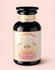 A decorative glass jar labeled "Tea for Abundance: Goddess Green: Apricot & Lychee Green Tea," featuring pastel art, floral accents, and gold details, contains a green tea blend with apricot, lychee, and organic lemongrass by Magic Hour. Known for its refreshing flavor and metabolism-supporting properties, the jar is displayed on a pale pink background.