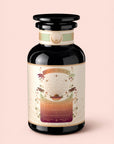 A dark glass jar labeled “Tea for Adventure: Horchata Chai: Spiced Yerba Mate” from Magic Hour, adorned with celestial and botanical illustrations—and a touch of Tulum’s spirit—on a cream-colored label, set against a light pink background.