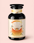 A dark glass jar labeled "Tea for Cleansing: Queen of the South: Herbal Cafe de Olla," adorned with decorative floral and celestial designs, sits against a pale pink background. This antioxidant-rich, caffeine-free Cocoa Detox Tea exudes an aura of purity and indulgence.