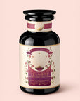 A dark glass jar with a gold and pink label reading "Tea for Love: Soulmate" from Magic Hour, adorned with stars, leaves, and sunburst designs, sits on a light pink background. This enchanting Black Tea with Chocolate & Raspberries blends hand-selected Fair Trade leaves and stunning artwork to celebrate every pour.