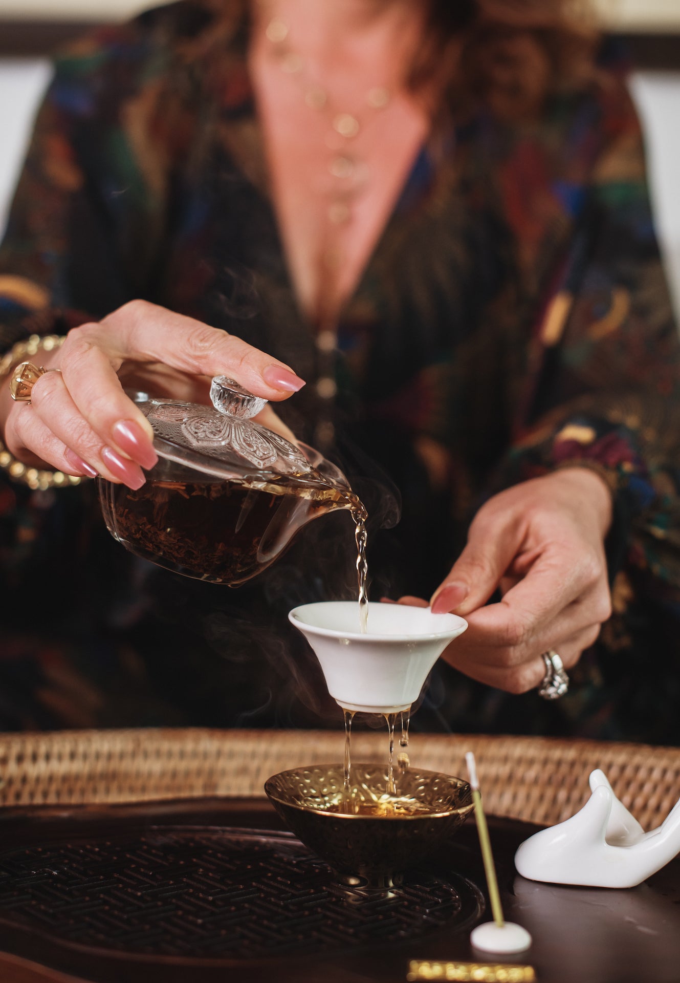 A person in a patterned blouse is gracefully pouring hot tea from a glass teapot into a white teacup, with wisps of steam rising. The scene includes a wicker tray, a small incense stick, and a white ceramic holder on a wooden table.
