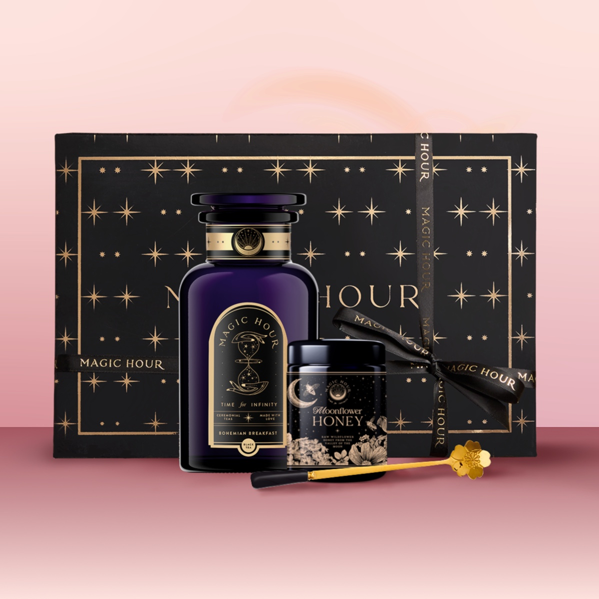 The Tea & Honey Gift Set features a dark blue bottle, wildflower honey with a golden wand, and a black box with gold stars and “Magic Hour”—the perfect touch to elevate your morning ritual on a soft pink background.
