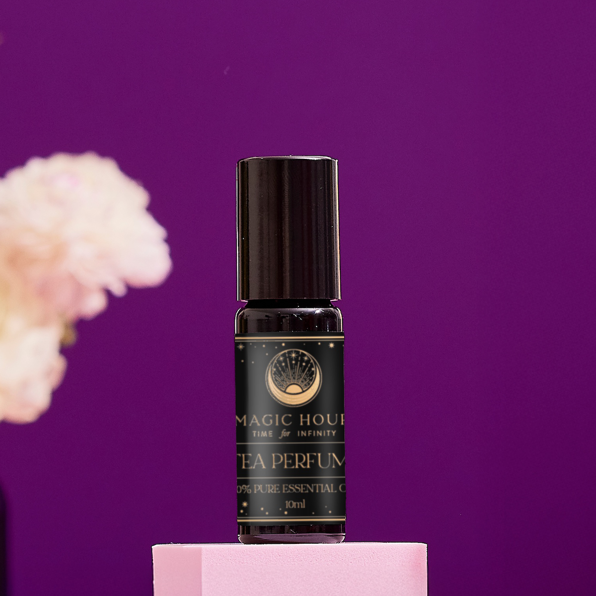 A small black bottle labeled "Tea Perfume Oil" rests on a pink pedestal against a purple background, with a blurred light-hued flower to the left, emphasizing this elegant natural fragrance.