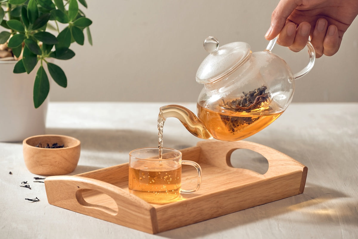A hand pouring tea from a glass teapot into a glass cup, both placed on a wooden tray. A bowl of tea leaves is nearby, and a green plant is in the background on a light-colored surface.