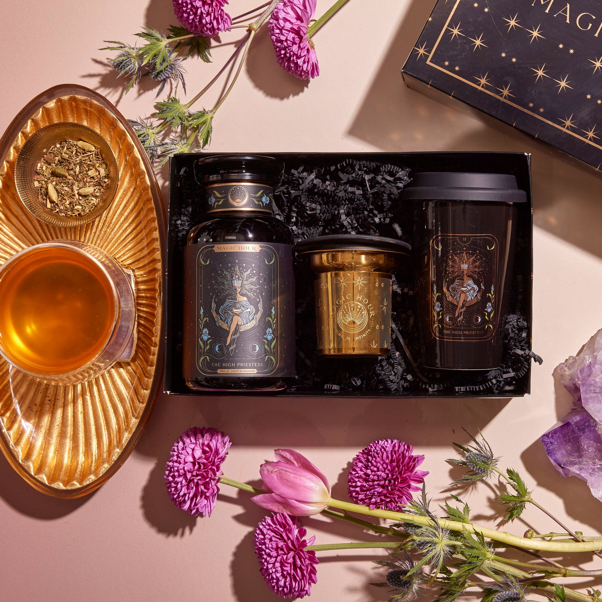 The Tea and Tarot to Go Gift Set presents a stunningly curated ensemble: it includes a jar, candle, and travel mug embellished with celestial artwork, encircled by purple blooms and an amethyst crystal. Complementing this mystical set is a golden cup of herbal tea on a matching saucer.