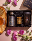 The Tea and Tarot to Go Gift Set presents a stunningly curated ensemble: it includes a jar, candle, and travel mug embellished with celestial artwork, encircled by purple blooms and an amethyst crystal. Complementing this mystical set is a golden cup of herbal tea on a matching saucer.