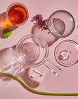 A Tea-in-Hand: The Perfect Steep Side-pour Ceremonial Teapot, two cups (one with a reddish liquid), flowers, and a glass stirrer are arranged artistically on a pink background. The shadows cast by the objects and flowers create a visually appealing composition, perfect for enjoying loose leaf teas.