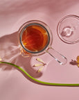 A glass cup filled with loose leaf tea sits on a light pink surface next to a Tea-in-Hand: The Perfect Steep Side-pour Ceremonial Teapot made of borosilicate glass by Espresso Parts. Two yellow daisies and their shadows are visible, one lying horizontally across the table and the other at an angle, adding a touch of nature to the setting.