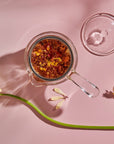 A top view of a borosilicate glass Espresso Parts Tea-in-Hand: The Perfect Steep Side-pour Ceremonial Teapot containing an infusion of loose leaf teas, positioned against a pale pink background. The teapot's lid is placed beside it. Two yellow daisies and a delicate dragonfly decoratively accompany the scene.