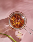A **Tea-in-Hand: The Perfect Steep Side-pour Ceremonial Teapot** by **Espresso Parts** made of borosilicate glass filled with floral loose leaf teas is placed on a pink surface with a daisy and a decorative model of a dragonfly nearby. The shadows of the daisy and dragonfly are visible, adding a whimsical feel to the scene.
