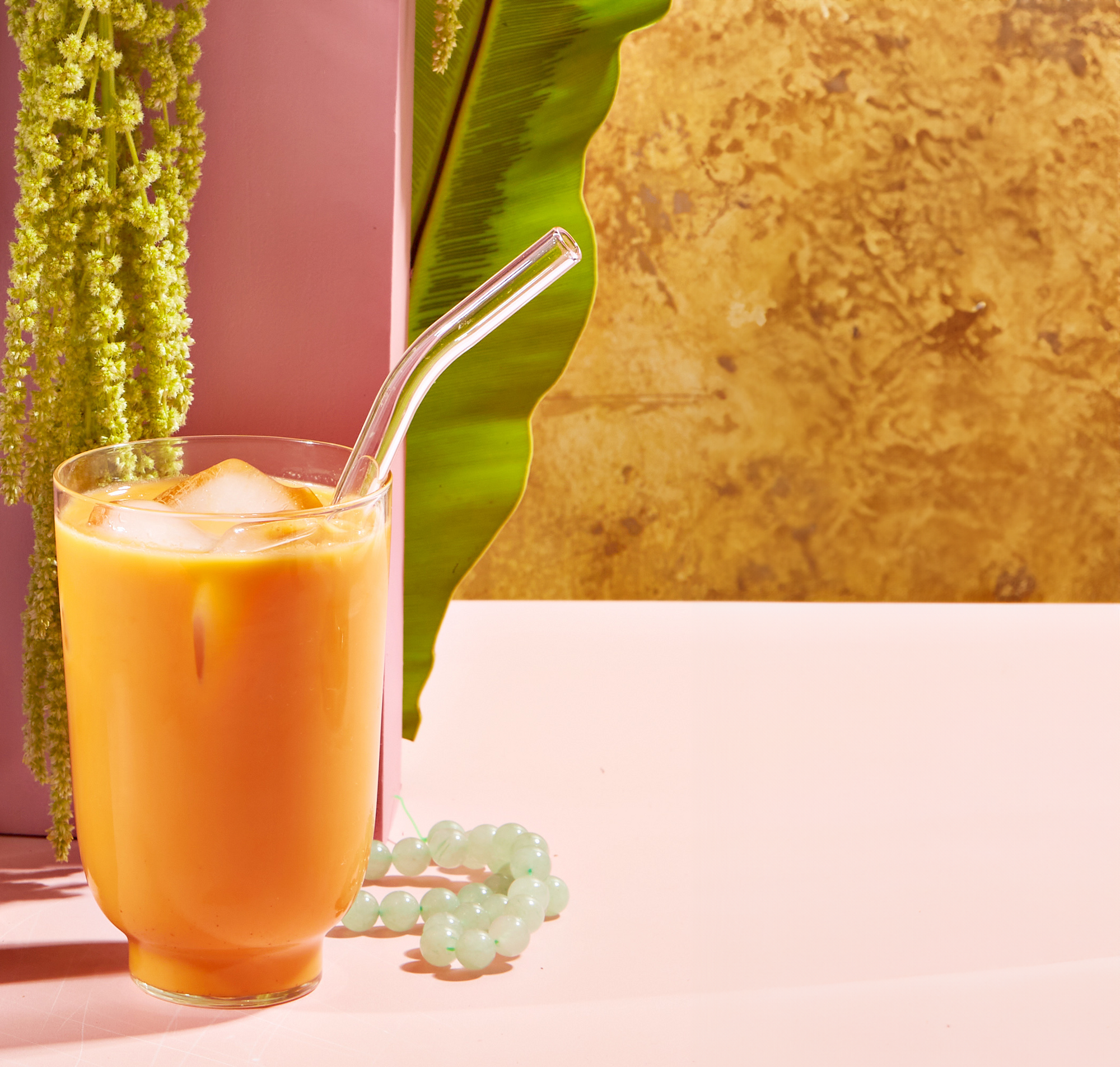 A glass of iced orange-colored drink with a transparent straw sits on a pink surface. Green foliage and a pink wall form the backdrop, and a beaded jade bracelet lies next to the glass. Part of the Wanderlust Sampler Tea Set, the scene exudes a tropical and refreshing vibe.