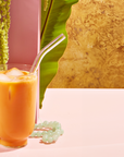 A glass of iced orange-colored drink with a transparent straw sits on a pink surface. Green foliage and a pink wall form the backdrop, and a beaded jade bracelet lies next to the glass. Part of the Wanderlust Sampler Tea Set, the scene exudes a tropical and refreshing vibe.