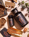 A flat lay of wellness products including jars of herbs, The Emperor: Green Chaga Chai with Lion's Mane and Reishi for immune support, a tin containing incense sticks, cinnamon sticks, dried flowers, and a box of mindful minutes cards. The items are beautifully arranged on a white surface with decorative gold and black paper underneath some pieces.