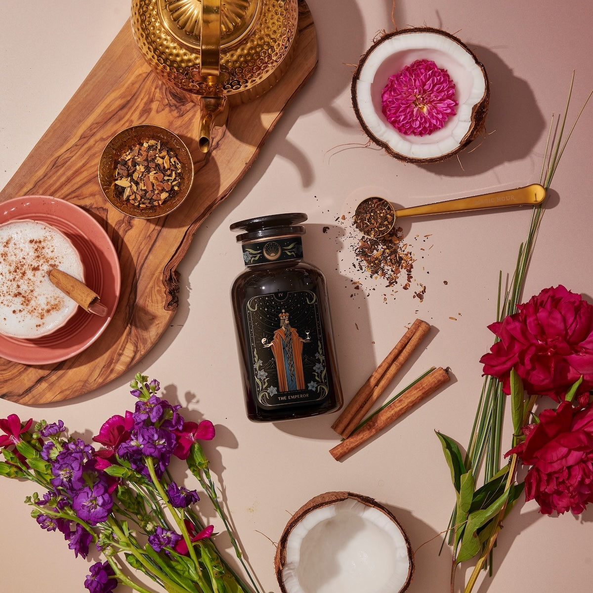 An artistic flat lay features a black perfume bottle with a floral design in the center, surrounded by vibrant flowers, cinnamon sticks, a halved coconut, a teapot, a bowl of tea leaves from the Monthly Magic First Sips Tea Subscription Box, a cup of tea with frothy milk, and a wooden tray.