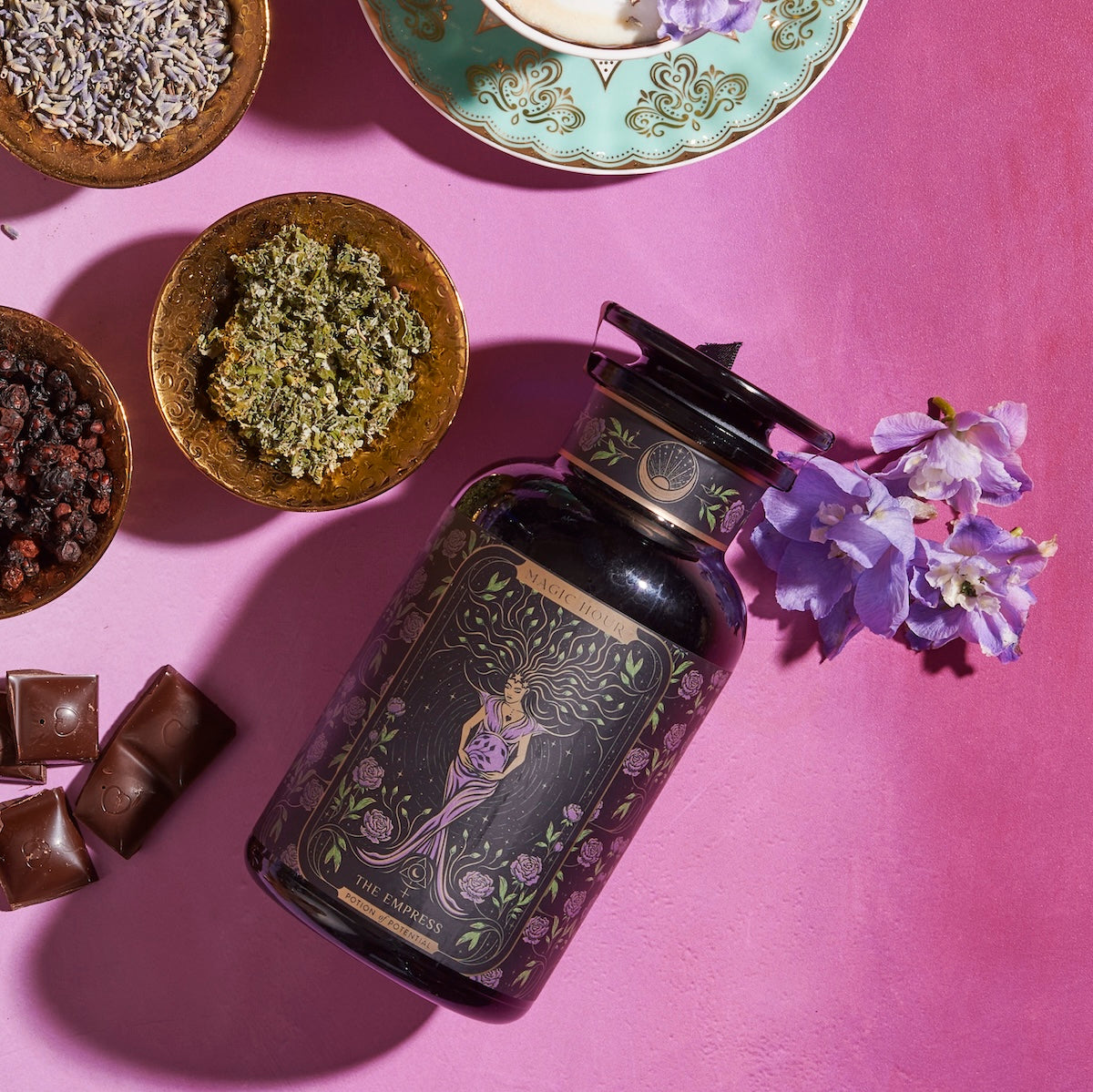 A jar adorned with an intricate design featuring a woman, labeled "THE EMPRESS," is surrounded by bowls containing dried herbs, lavender, raisins, and chocolates. A bouquet of purple flowers is placed beside it on a pink surface, capturing the essence of the Magic Hour's Tea and Tarot to Go Gift Set.