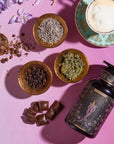 A flat lay of The Empress: Lavender Currant Shatavari Cocoa Tea of Nurturing Creativity by Magic Hour in an ornate cup and saucer, surrounded by dried flowers, organic herbs, chocolate pieces, and a decorative jar on a pink and purple gradient background. A sprig of fresh purple flowers and a white calla lily are also in the scene.