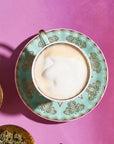 A cup of cappuccino with frothy milk on top is placed on a decorative turquoise saucer with intricate patterns. The saucer rests on a pink surface. Surrounding the cup are small bowls containing organic herbs, creating an inviting atmosphere for a Magic Hour: The Empress: Lavender Currant Shatavari Cocoa Tea of Nurturing Creativity experience.