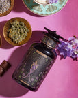 A jar adorned with an intricate design featuring a woman, labeled "THE EMPRESS," is surrounded by bowls containing dried herbs, lavender, raisins, and chocolates. A bouquet of purple flowers is placed beside it on a pink surface, capturing the essence of the Magic Hour's Tea and Tarot to Go Gift Set.