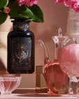 A decorative arrangement featuring a dark glass bottle with ornate design, a clear glass jug of Magic Hour's The High Priestess: Wisdom Tea for Powerful Dreams & Illuminated Insights, a pink crystal sphere on a stand, a white crystal tower, a black crystal obelisk, and a bouquet of pink flowers in a vase, all on a pink surface.