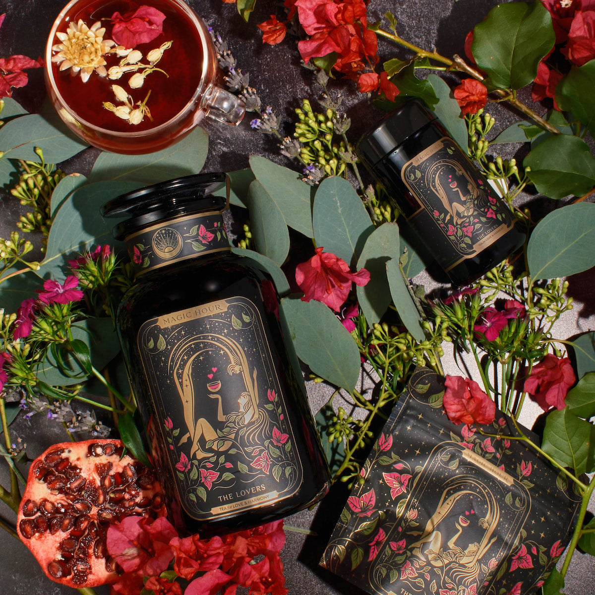 An opulent flat lay image showcases a large black apothecary-style bottle labeled "The Lovers" from The Lovers Box, accompanied by a smaller matching bottle. A cup of tea from the Magic Hour Branded Gift Box, garnished elegantly, sits nearby. An open pomegranate and an array of scattered red and purple flowers with lush green leaves complete the arrangement on a dark surface.