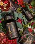 An opulent flat lay image showcases a large black apothecary-style bottle labeled "The Lovers" from The Lovers Box, accompanied by a smaller matching bottle. A cup of tea from the Magic Hour Branded Gift Box, garnished elegantly, sits nearby. An open pomegranate and an array of scattered red and purple flowers with lush green leaves complete the arrangement on a dark surface.