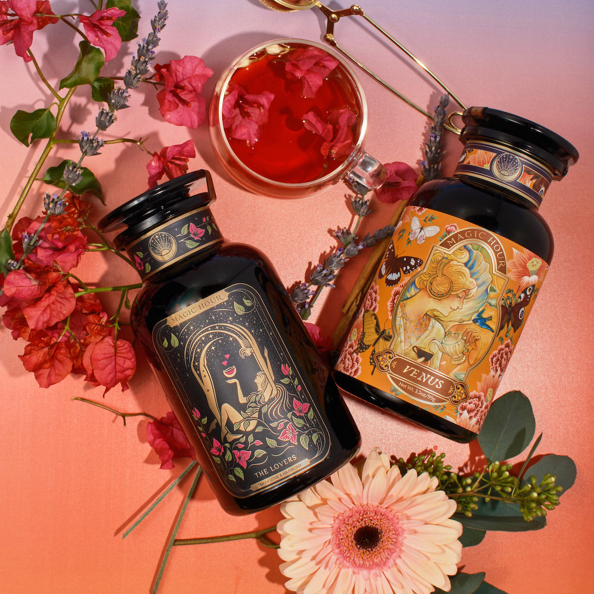 Two ornate black bottles of herbal supplements, labeled "The Lovers" and "Venus," lie flat on a pink-purple gradient background. A glass cup from Magic Hour's The Lovers Bundle with Venus + The Lovers Tea, featuring a beautifying herbal blend with sensual flavors, floats beside them, surrounded by flowers and greenery, including a gerbera daisy and lavender.