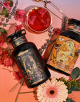 Two ornate black bottles of herbal supplements, labeled "The Lovers" and "Venus," lie flat on a pink-purple gradient background. A glass cup from Magic Hour's The Lovers Bundle with Venus + The Lovers Tea, featuring a beautifying herbal blend with sensual flavors, floats beside them, surrounded by flowers and greenery, including a gerbera daisy and lavender.