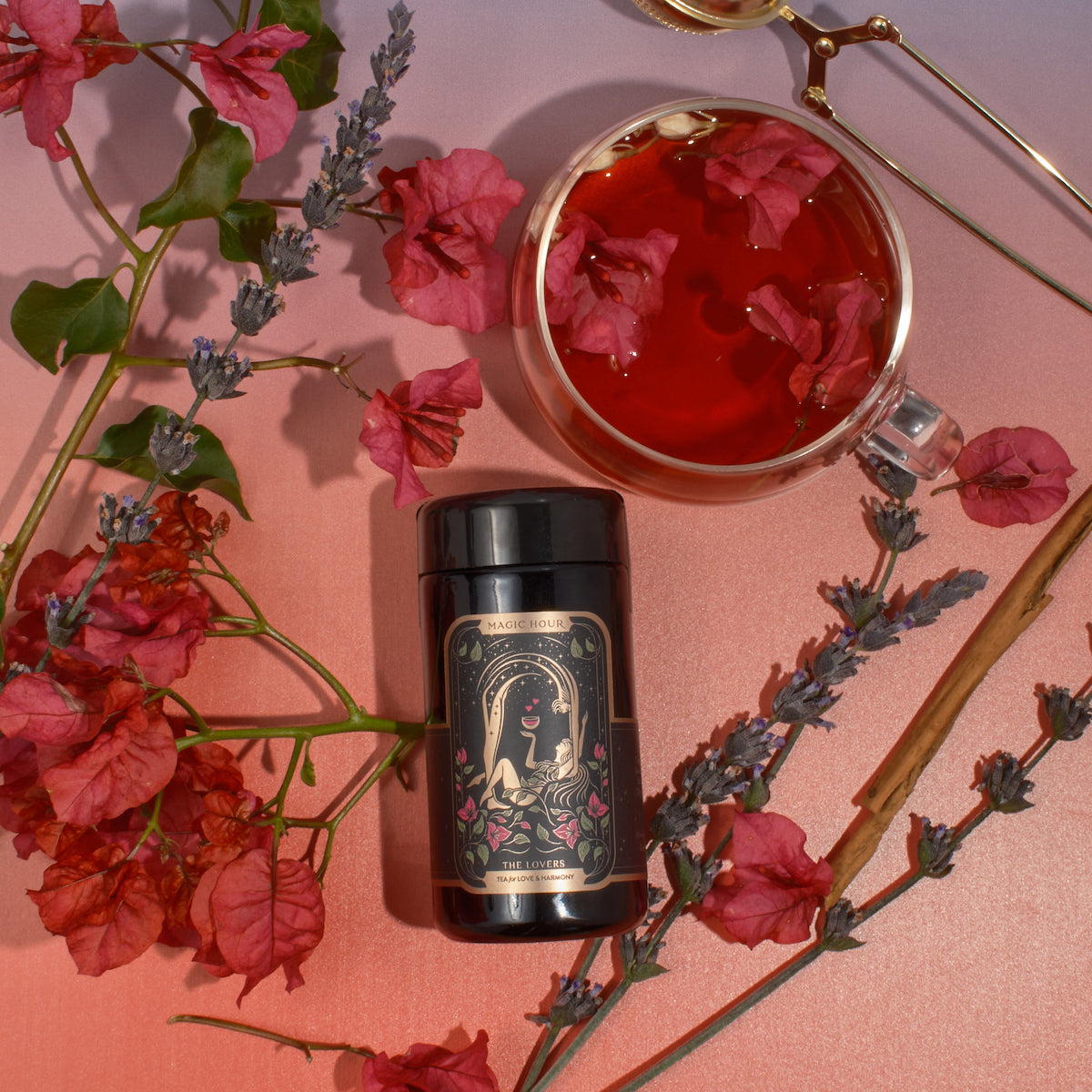 A cylindrical tin from The Lovers Box, adorned with illustrated roses and golden accents, is encircled by vibrant red and pink bougainvillea flowers, aromatic lavender sprigs, and cinnamon sticks. Close by, a glass cup of reddish tea garnished with bougainvillea petals rests on a gradient pink surface.
